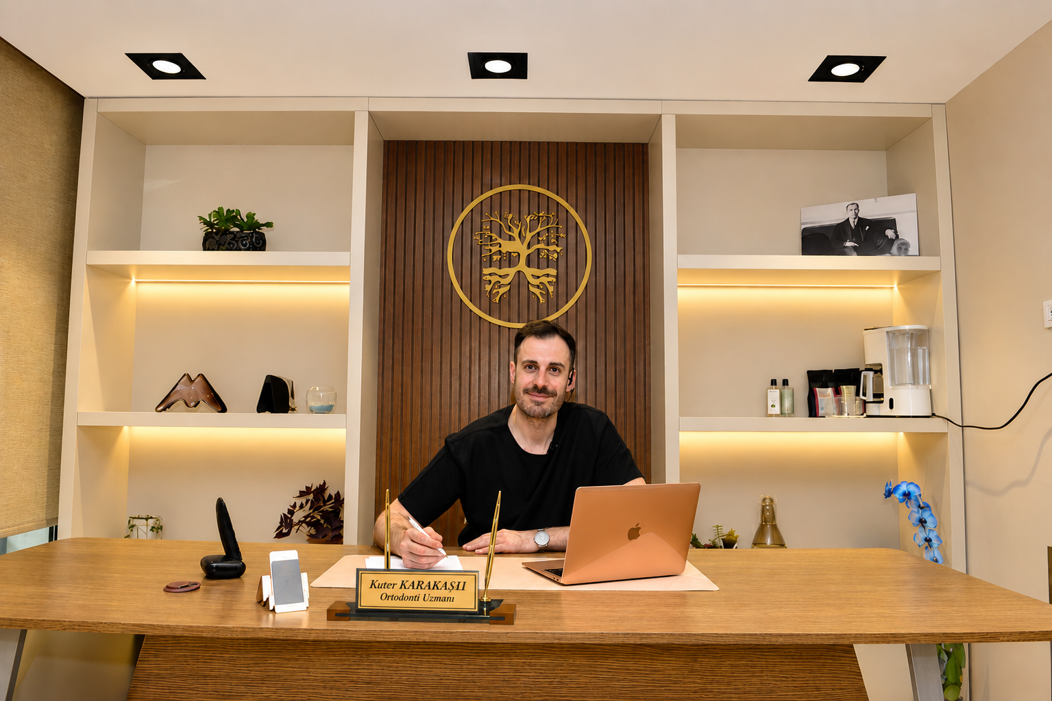 Dr. Kuter Karakaşlı, 4K Dent logosunun önünde ofis masasında